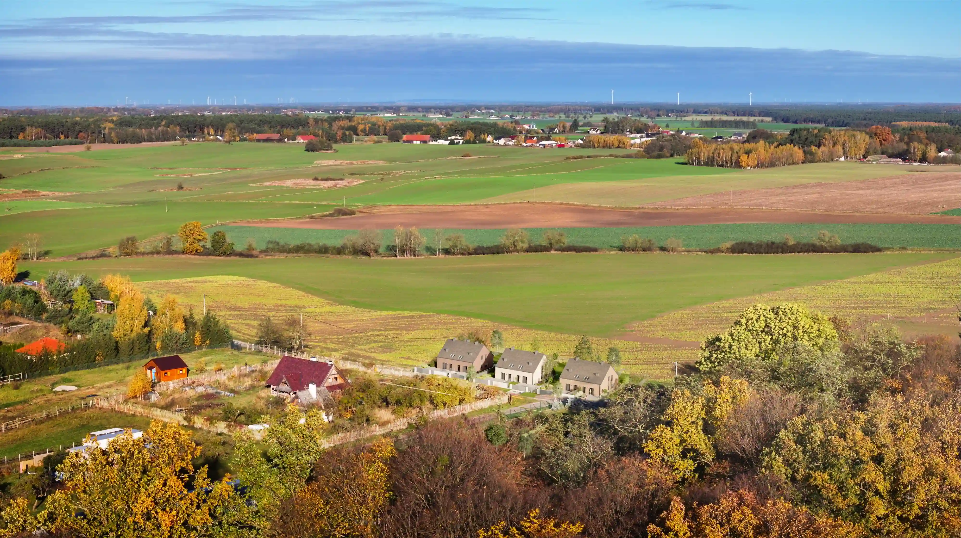 Widok z lotu ptaka na wiejski krajobraz w Potrzanowie. W dolnej części zdjęcia widać kilka domów otoczonych drzewami i ogrodami. W tle rozciągają się szerokie pola uprawne i łąki, które tworzą mozaikę różnokolorowych połaci ziemi. Jesienne kolory drzew kontrastują z zielenią pól, a w oddali widoczne są inne gospodarstwa i lasy.